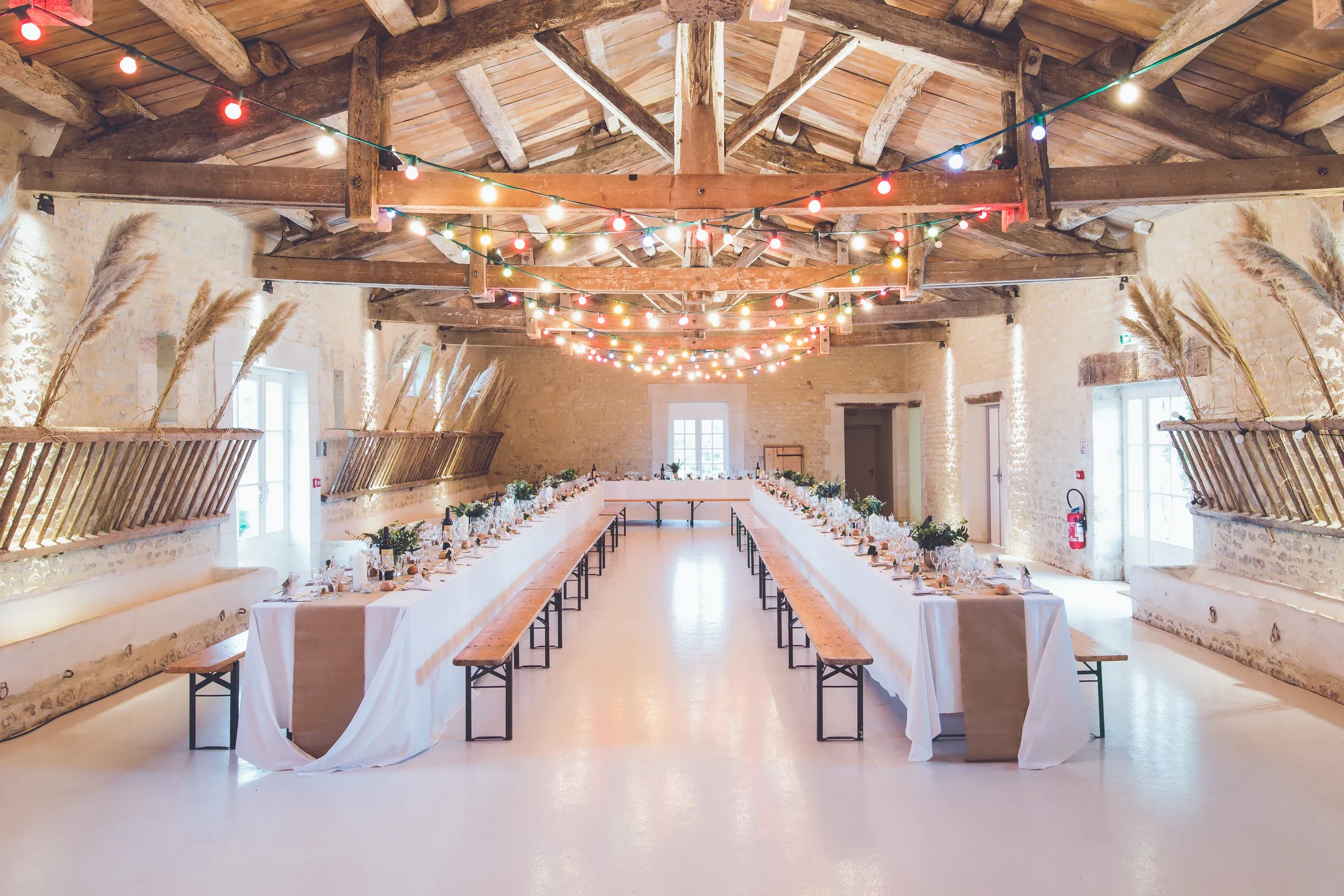 Long tables and timber benches set for dinner in a stone hall under festoon lights and exposed roof beams