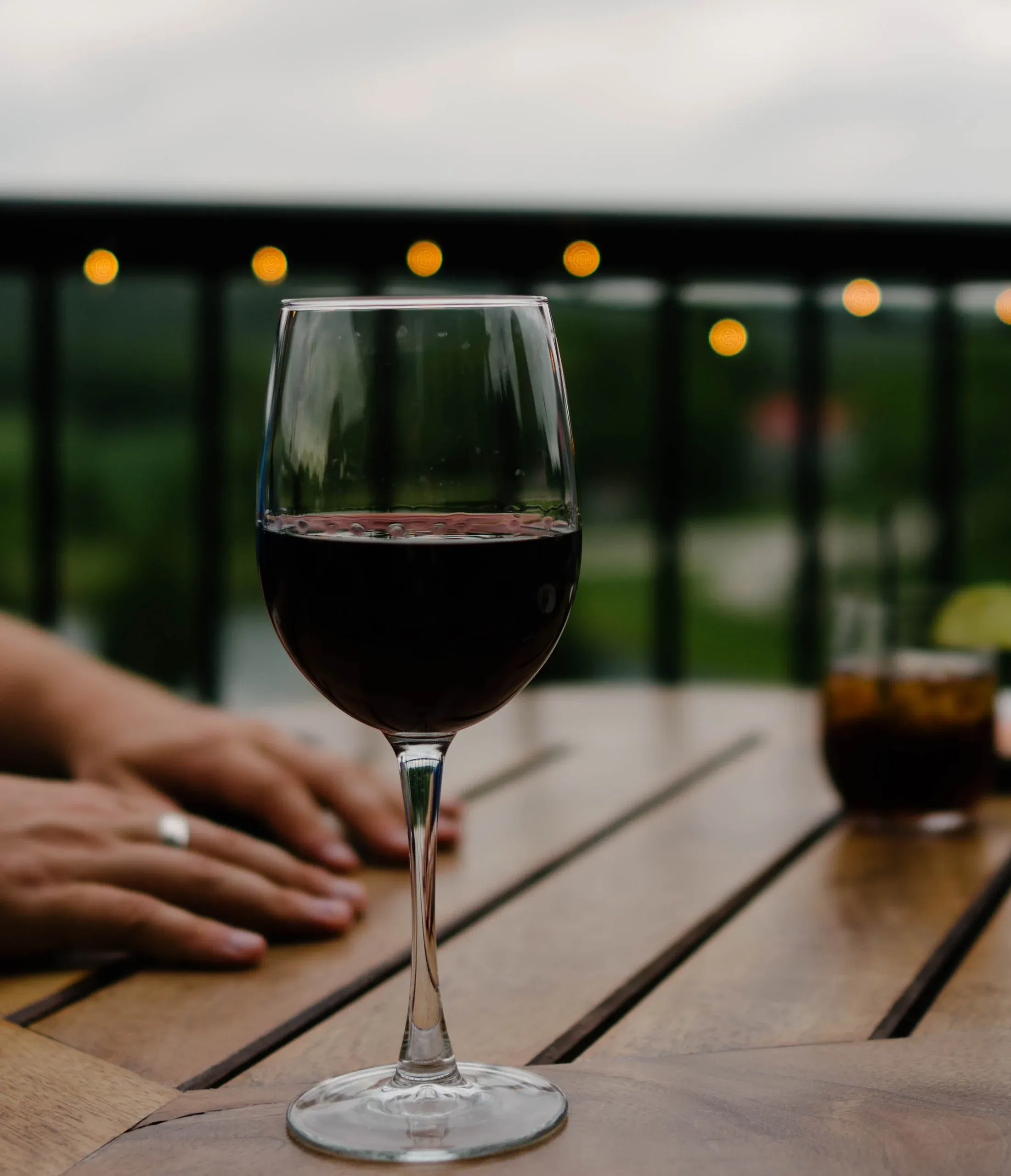 A glass of red wine on an outdoor timber table at dusk, with festoon lights glowing along the rail behind