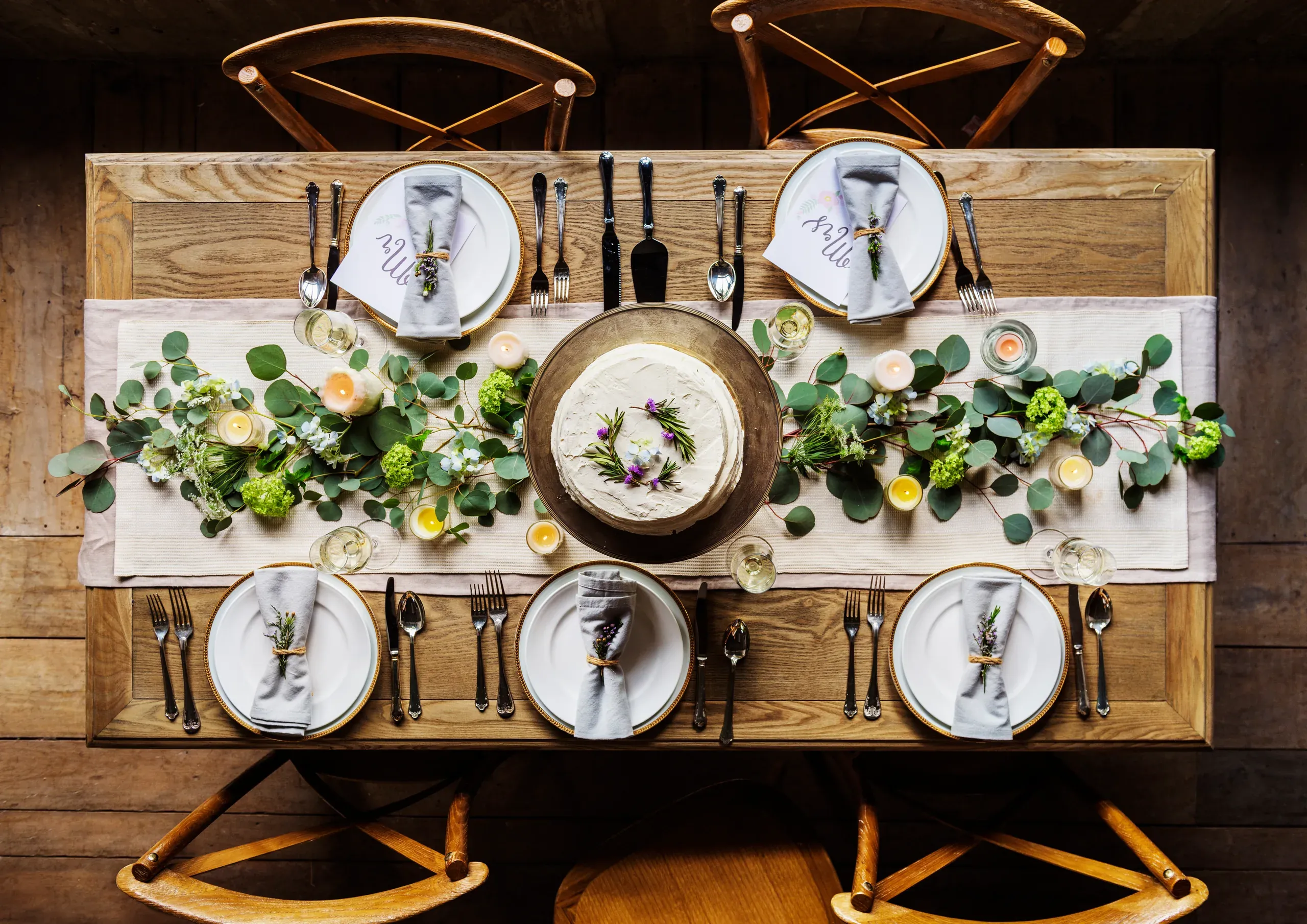 A timber table seen from above, set with plates, a cake and a runner of eucalyptus and small flowers