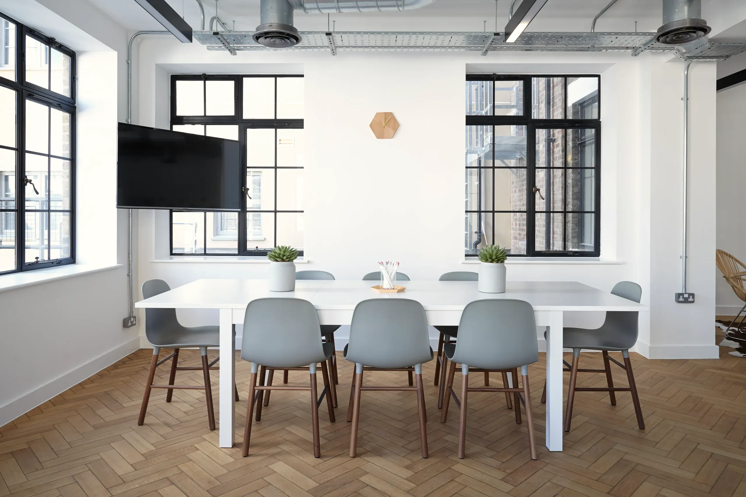 A bright meeting room with a wall screen, a long white table and grey chairs on a parquet floor