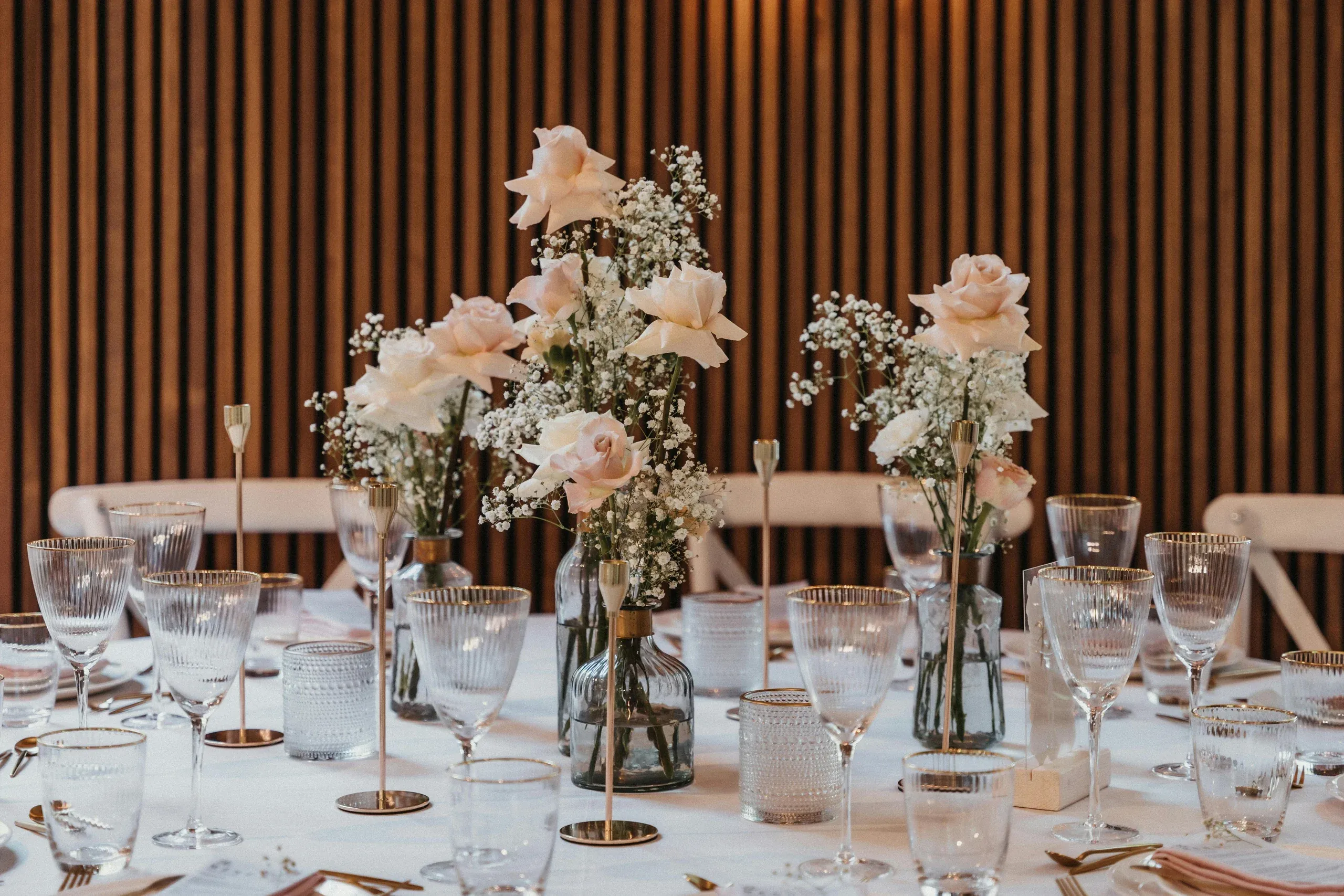 Blush roses and baby's breath in glass bottles on a dressed table before a slatted timber wall