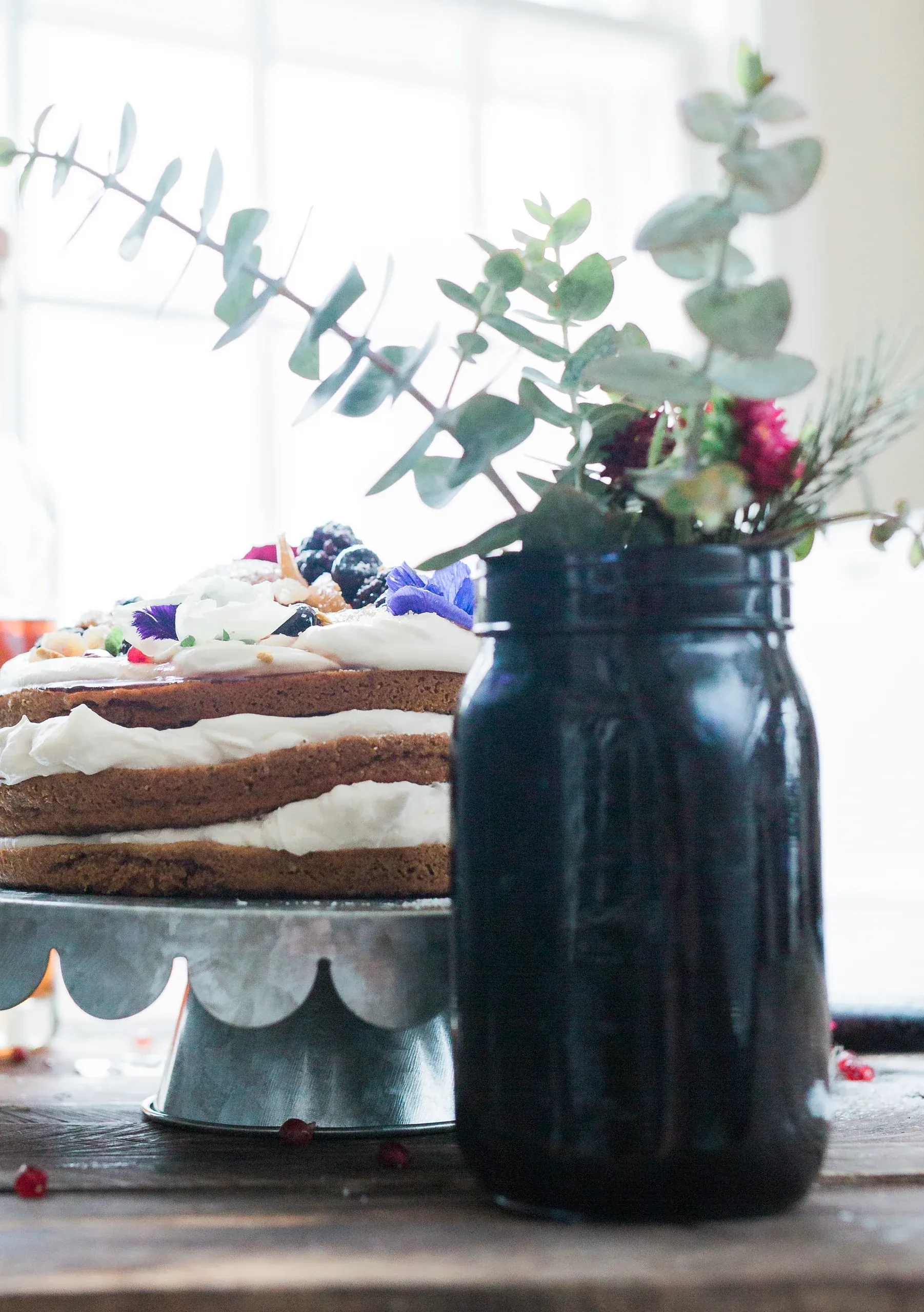 A layered naked cake with cream, berries and edible flowers beside a jar of eucalyptus in window light